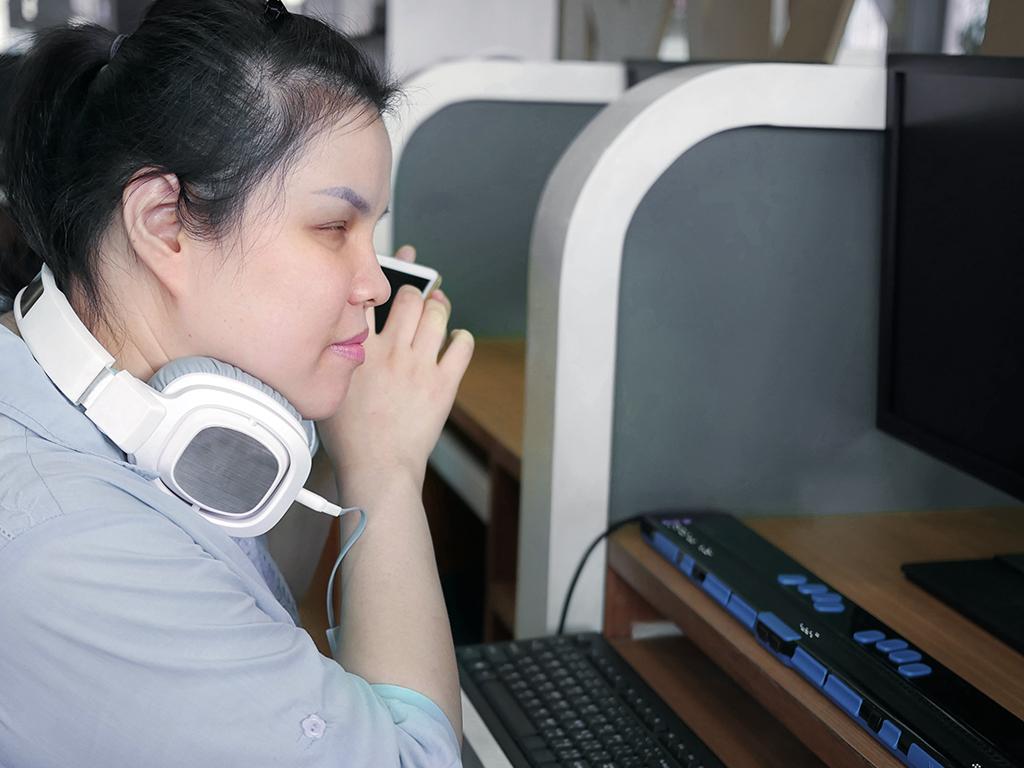 Blinde Frau am Computerarbeitsplatz mit Braille-Tastatur und Smartphone mit Voice Assistent
